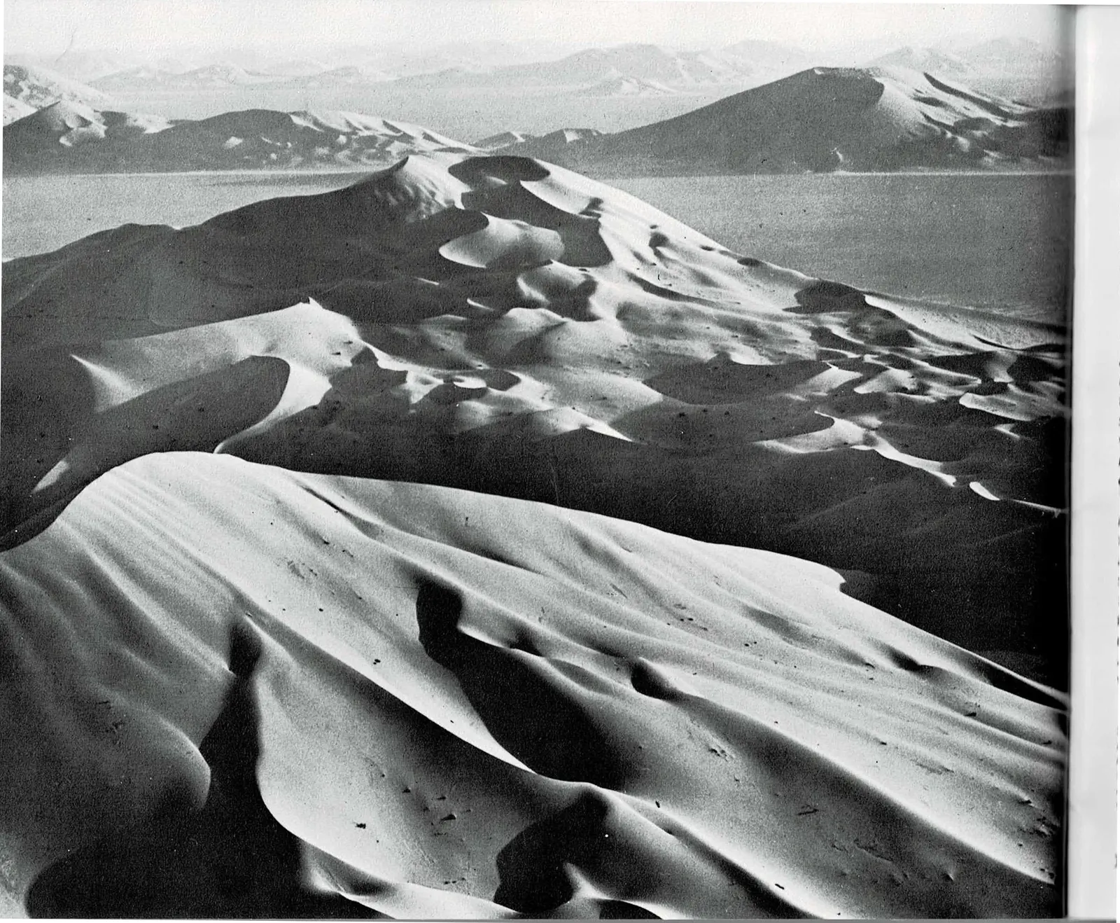 A two-page spread: at left, a full-bleed black and white aerial photograph of towering sand dunes in Saudi Arabia's Empty Quarter, their crests lit and rippled against a hazy desert horizon; at right, a text column recounting the 1872 Powell Survey's visit to Zion and quoting geologist Clarence Dutton's description of the Temples and Towers of the Virgin, beside a black and white photograph of dark, water-carved potholes and narrow sandstone walls.