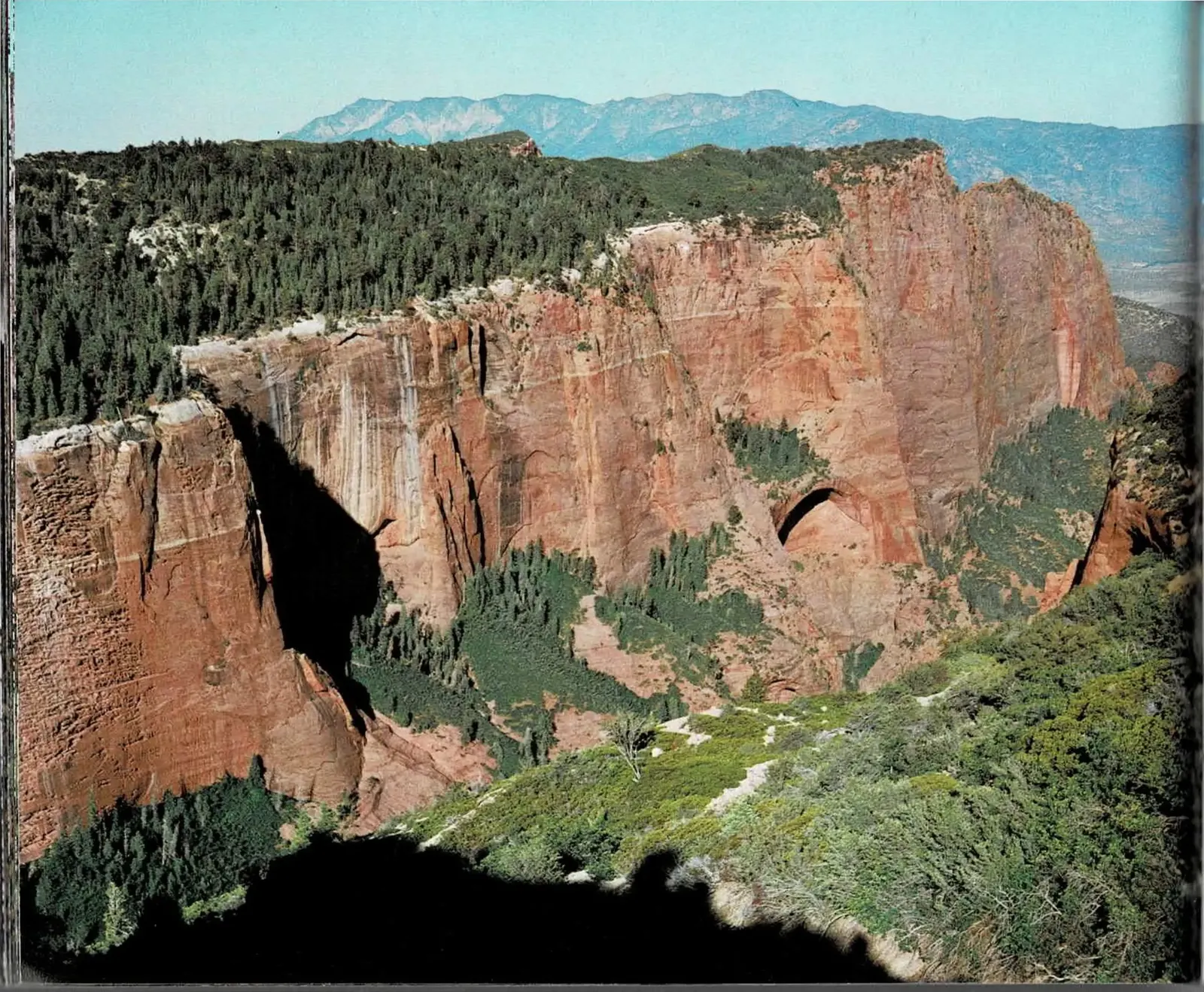 A two-page spread: at left, a full-bleed color photograph of a red-rock finger canyon in the Kolob, cliffs rising above forested slopes with a natural arch or alcove visible partway up the rock face and blue-hazed mountains receding into the distance; at right, a text column continuing mid-sentence, paired with a black and white photograph of a steep, crossbedded sandstone canyon dusted with snow and framed by conifers along its rim.