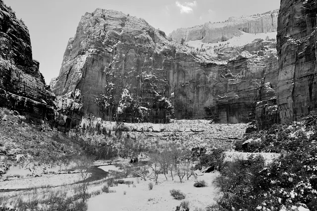A wide snow-dusted canyon under a tall sandstone wall, in black and white