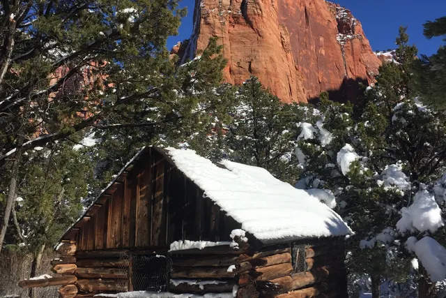 A small snow-roofed wooden cabin below a red sandstone monolith and evergreens